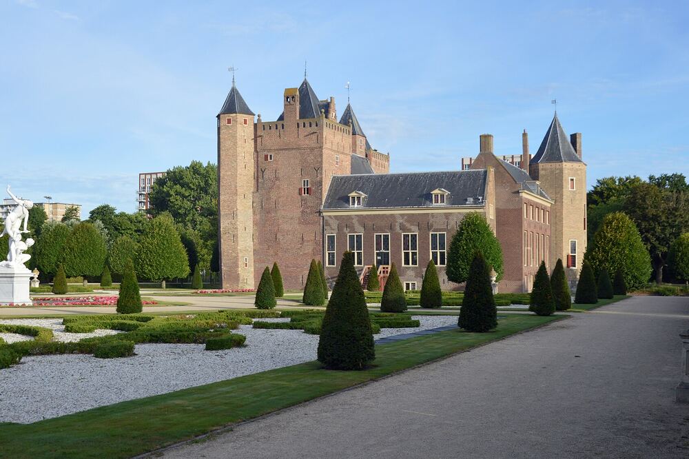 Kasteel Assumburg in Heemskerk met hoektorens en spitse daken, gezien over de symmetrische kasteeltuin met buxushagen, een wit tuinbeeld en een grindpad, onder een blauwe lucht